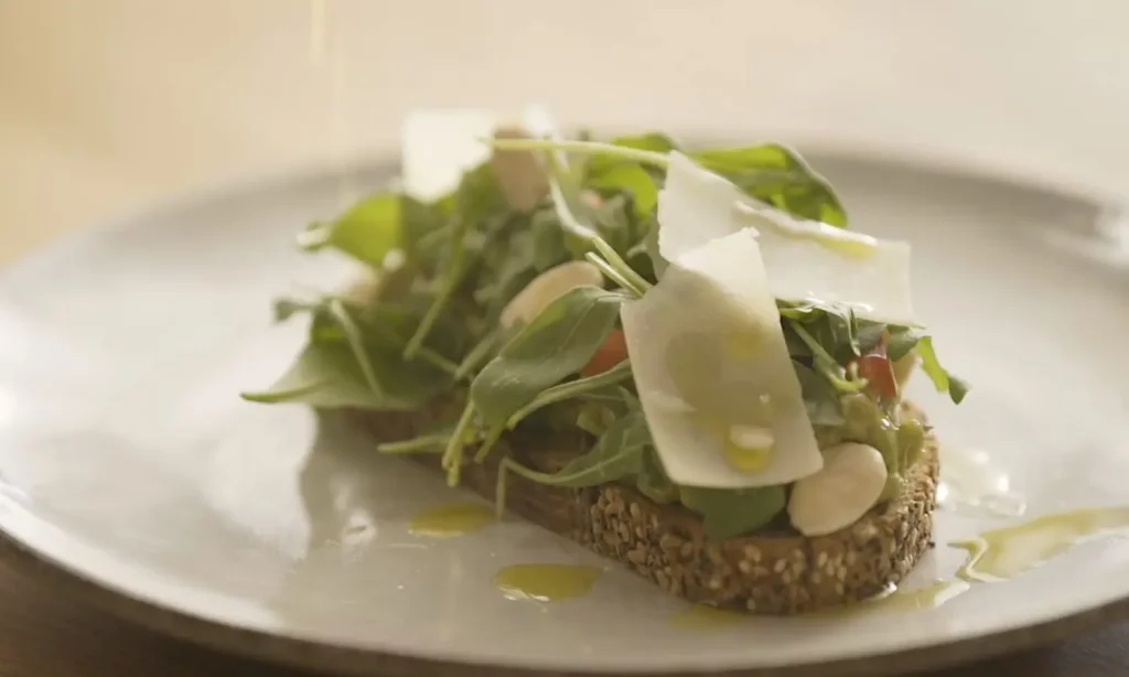 Tostada con aguacate, parmesano, rúcula y almendras sobre plato blanco